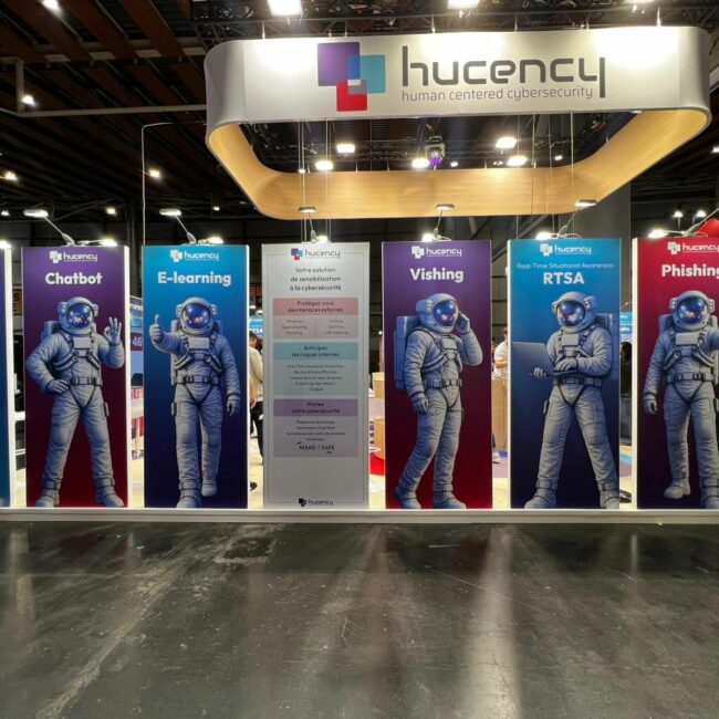 Estande da Hucency no Fórum InCyber, em Lille. Sete painéis coloridos apresentam Thomas, o CISO virtual representado como um astronauta. Ele ilustra a gestão conjunta da estratégia cibernética (phishing, e-learning, vishing) ao lado dos CISOs e dos diretores de TI, sob o lema «human centered cybersecurity».